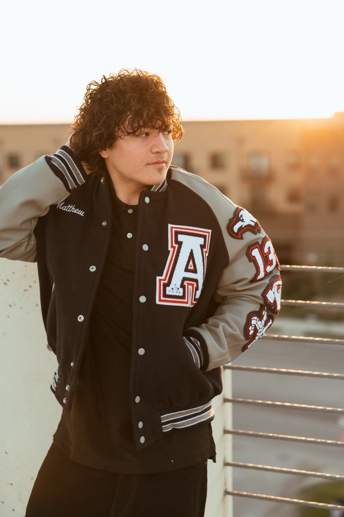 A person wearing a black and grey varsity letterman jacket poses at sunset against an urban railing.