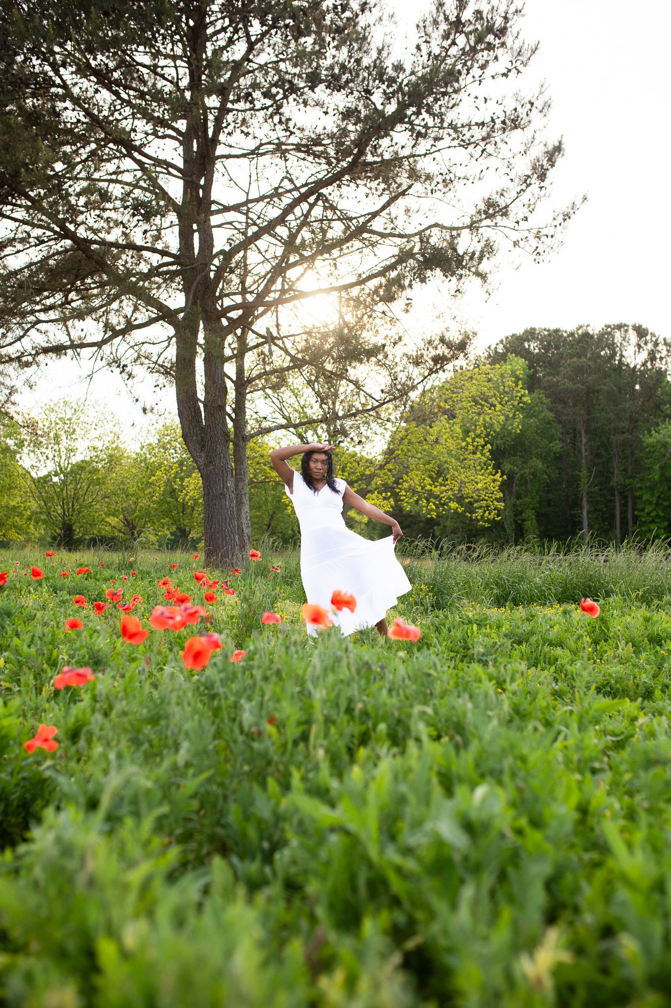A Woman in a flowing white dress twirls in a meadow with bright red poppies and a tall pine tree at sunset.