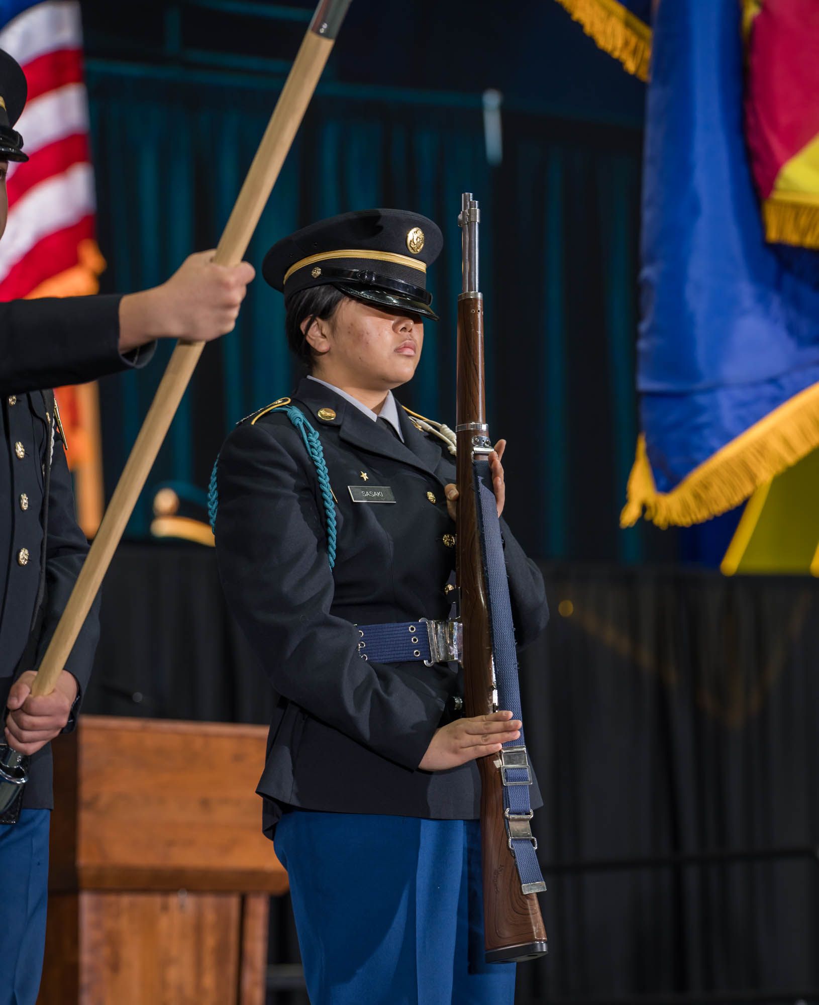 Honor guard member in dress uniform stands at attention holding a rifle during ceremonial event with flags in background.