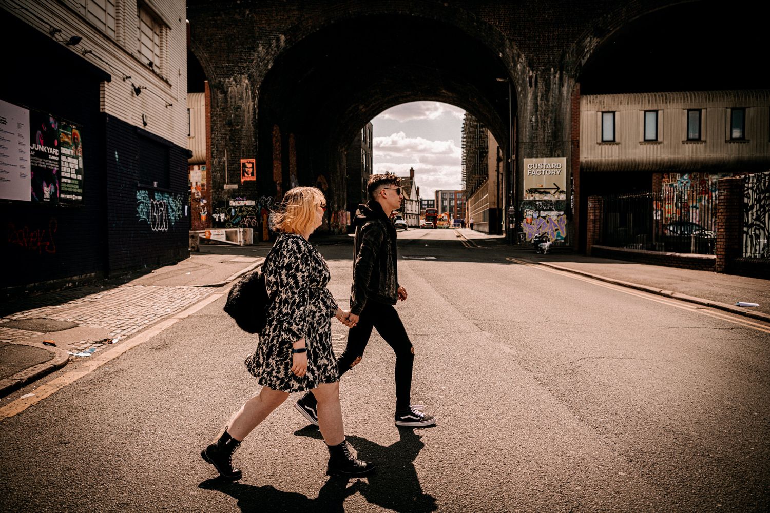 Two people walk beneath an urban railway arch on a sunny day casting long shadows on the street.