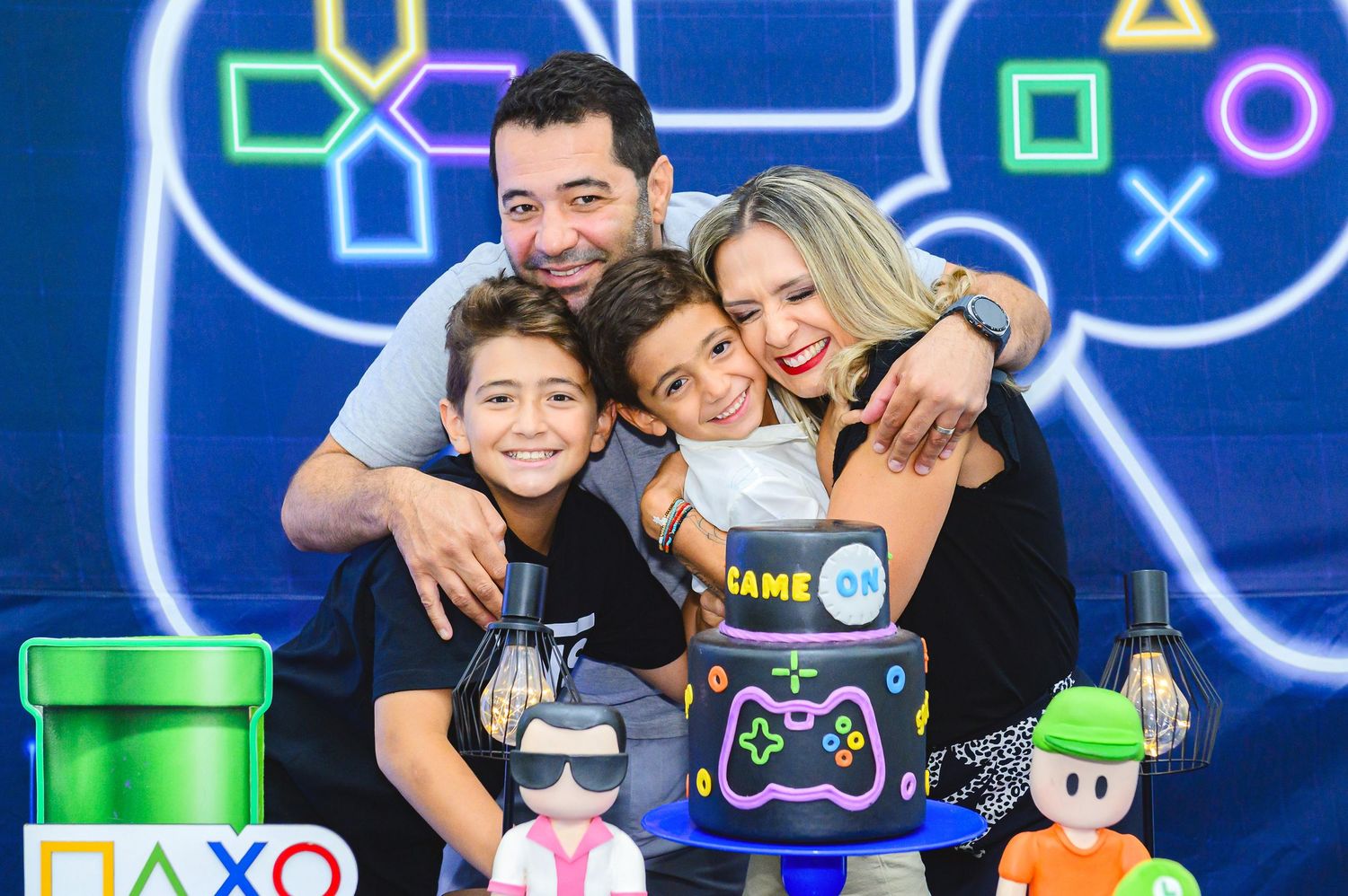A family poses together against a vibrant gaming-themed backdrop with neon PlayStation icons and gaming decorations.