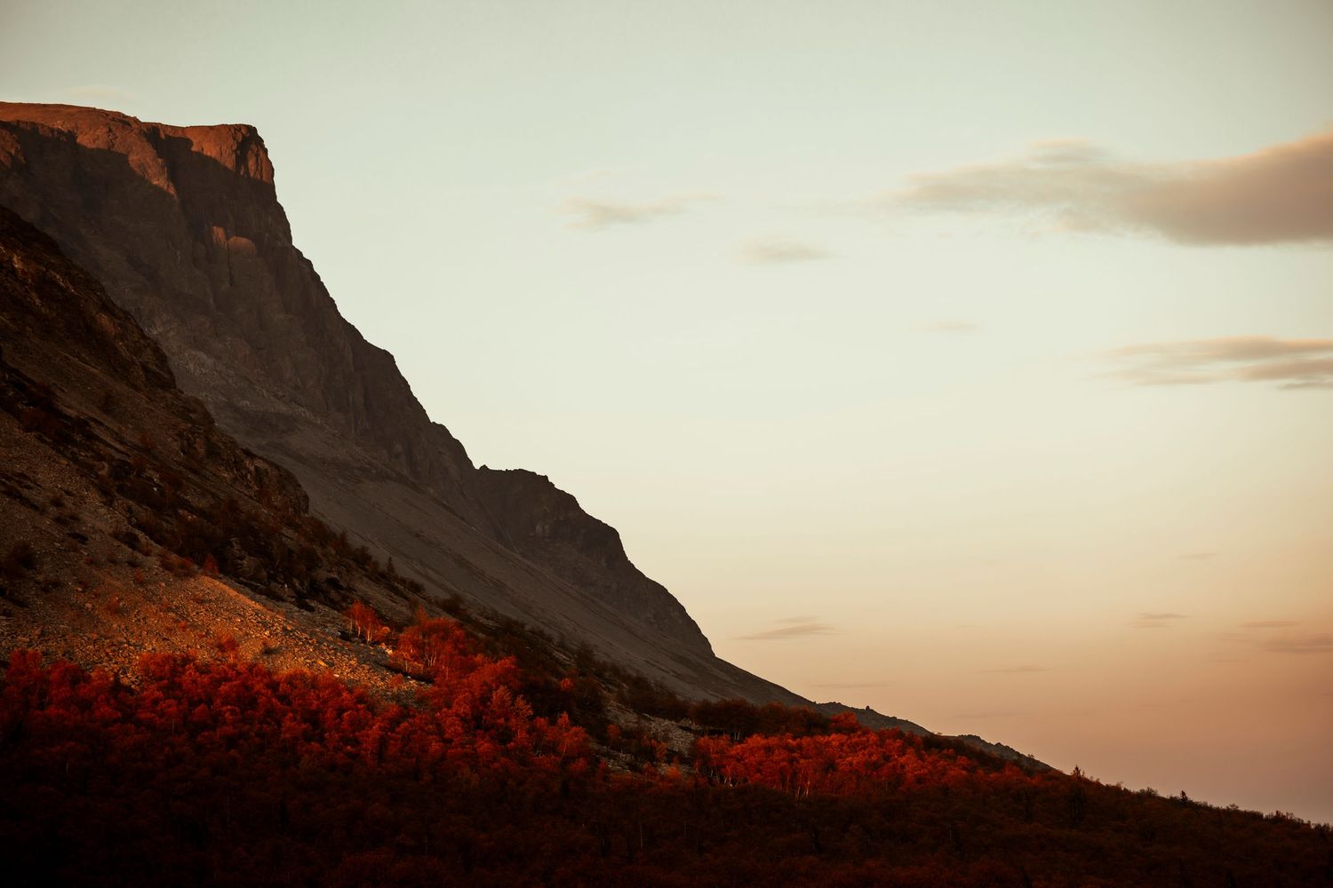 Dramatic mountain cliff silhouetted against sunset sky with red autumn foliage in foreground.