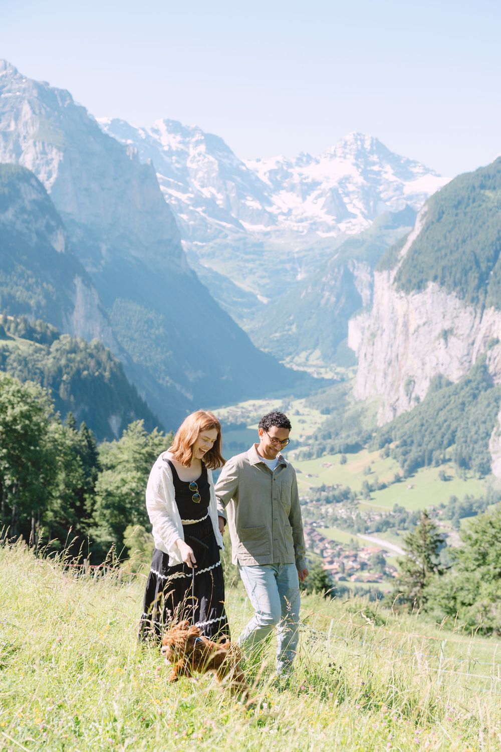 Two people walk together on a scenic mountain meadow with snow-capped Alpine peaks and a valley lake in the background.