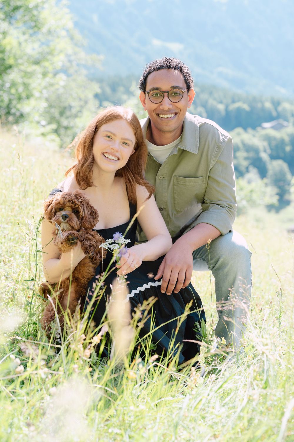 A couple sits lovingly together in a sunlit meadow of tall grass with mountains visible in the background.