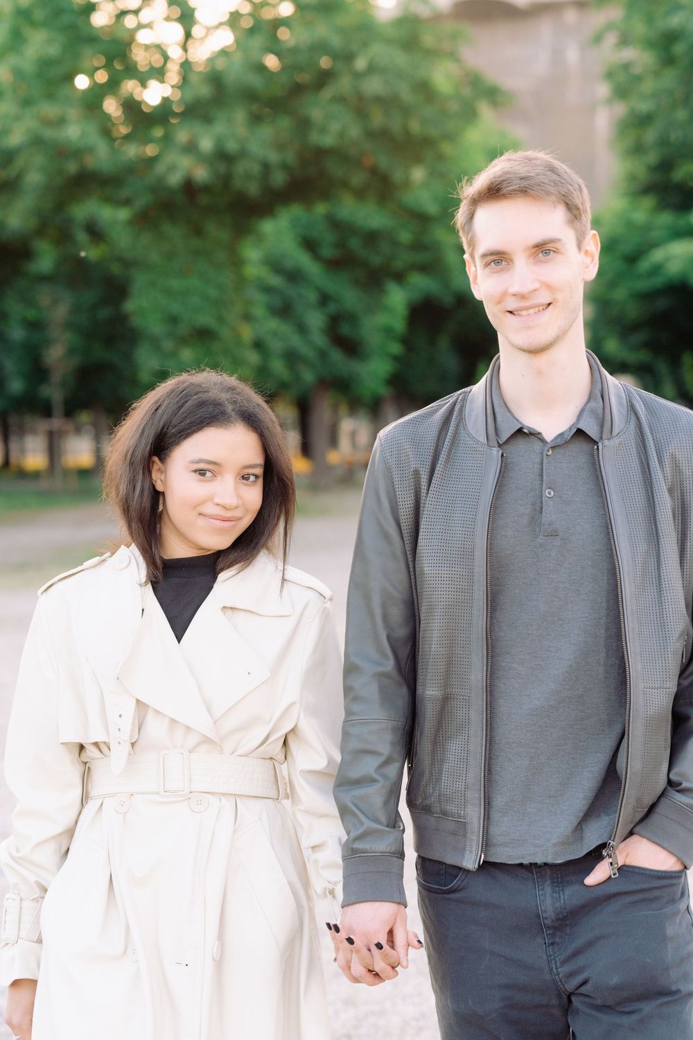 A couple holding hands outdoors with green trees and bokeh lights in the background.