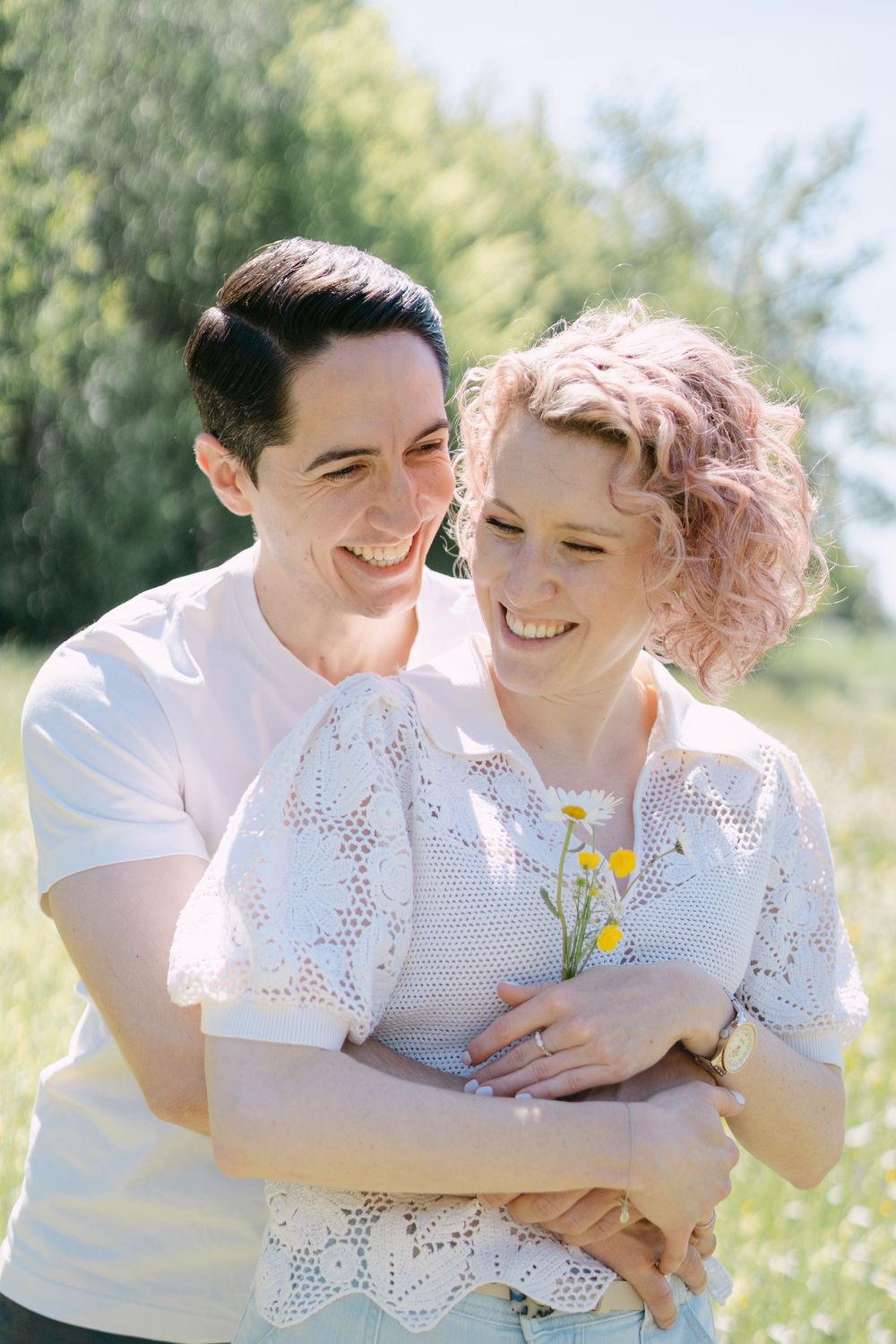 A couple laughing together in a sunny meadow with wildflowers, wearing white tops on a bright spring day.