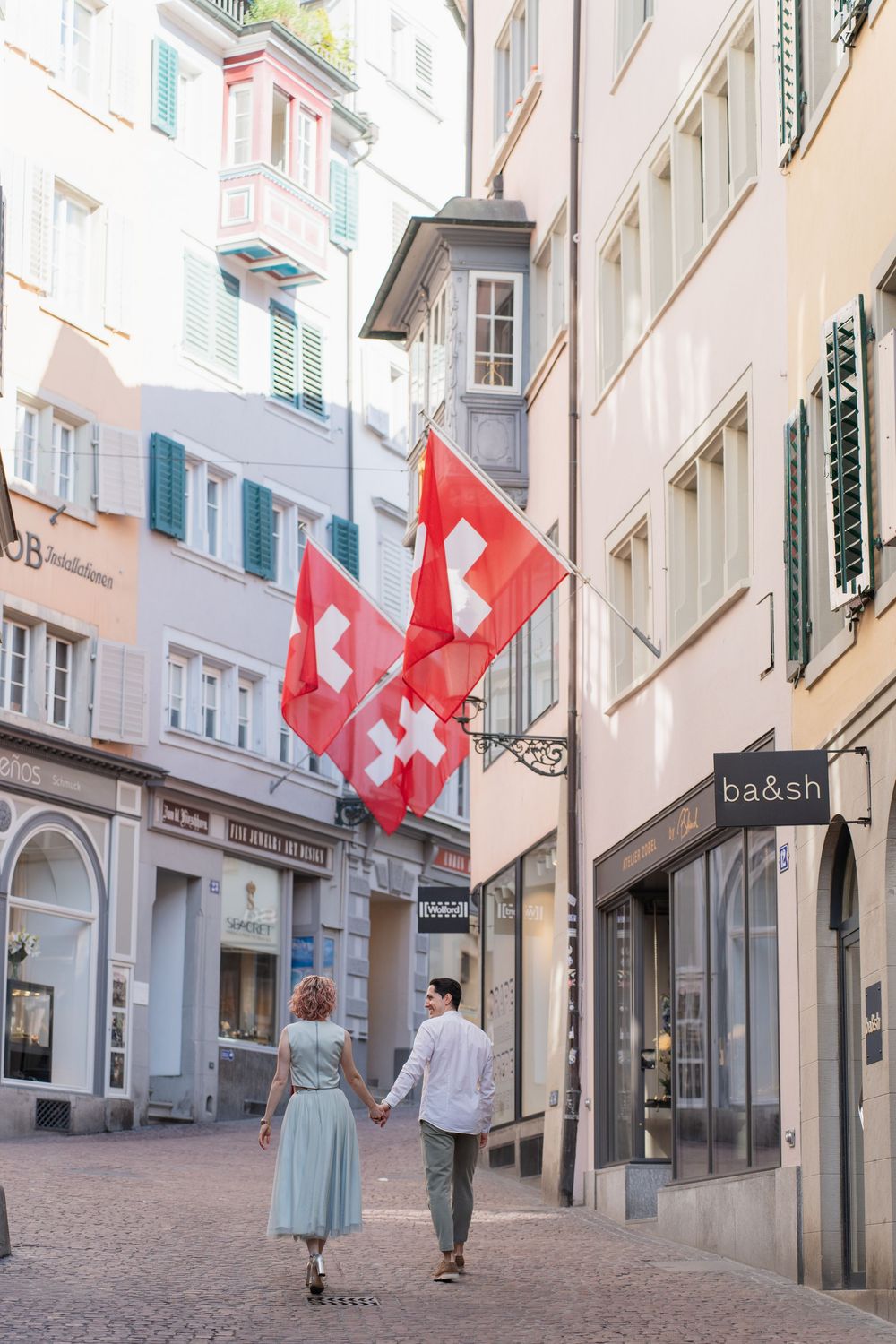 Couple walking down charming cobblestone street with Swiss flags hanging from historic buildings in Zurich, Switzerland.