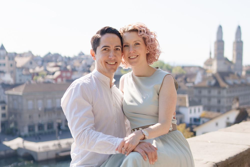A romantic couple smiles together on a rooftop overlooking a historic European city skyline with church spires.