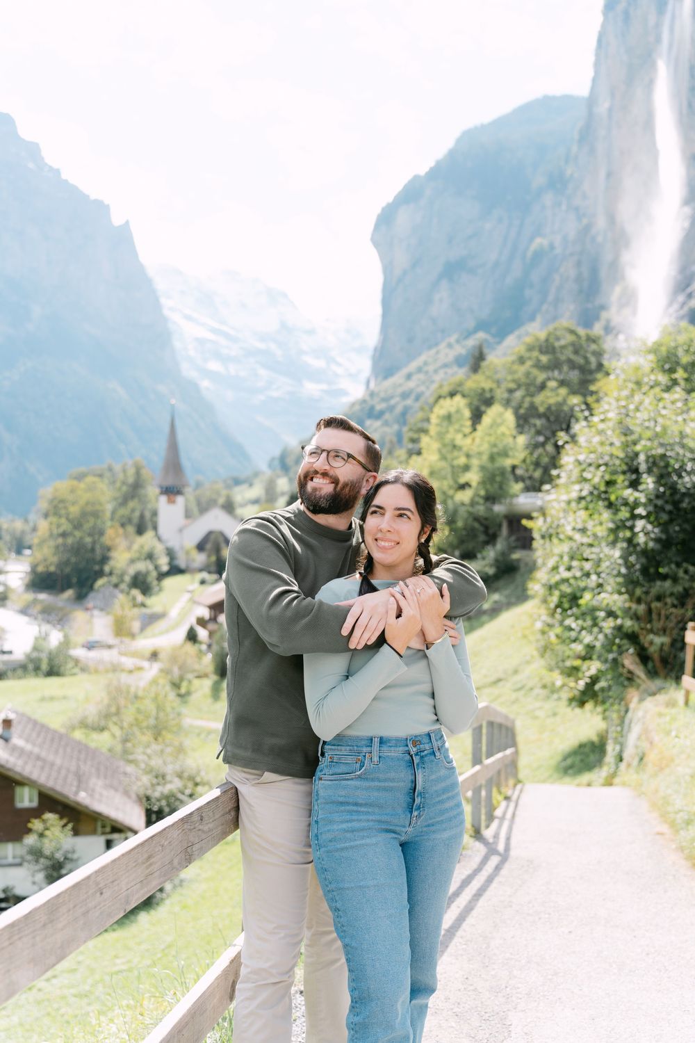 A couple embraces while standing on a path with dramatic Swiss Alps mountains and a waterfall in the background.