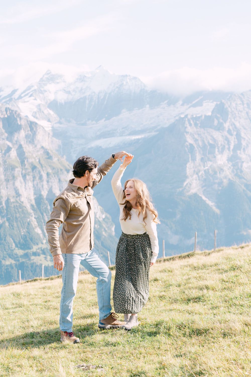 A couple dances joyfully on a grassy mountainside with stunning snow-capped Alpine peaks stretching across the horizon.