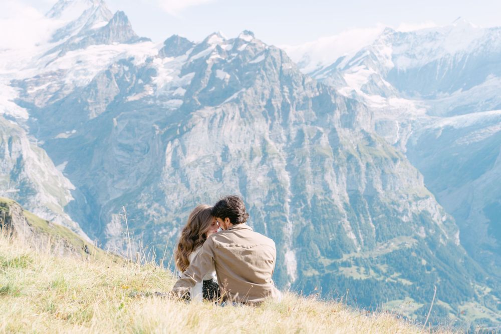 Someone sitting on a grassy hillside overlooking dramatic snow-capped mountain peaks in the Alps.