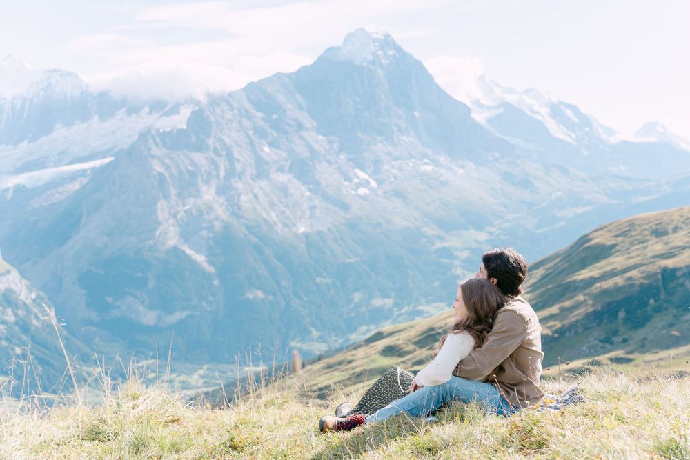 Someone sits on a grassy hillside overlooking majestic snow-capped mountain peaks in a serene alpine landscape.