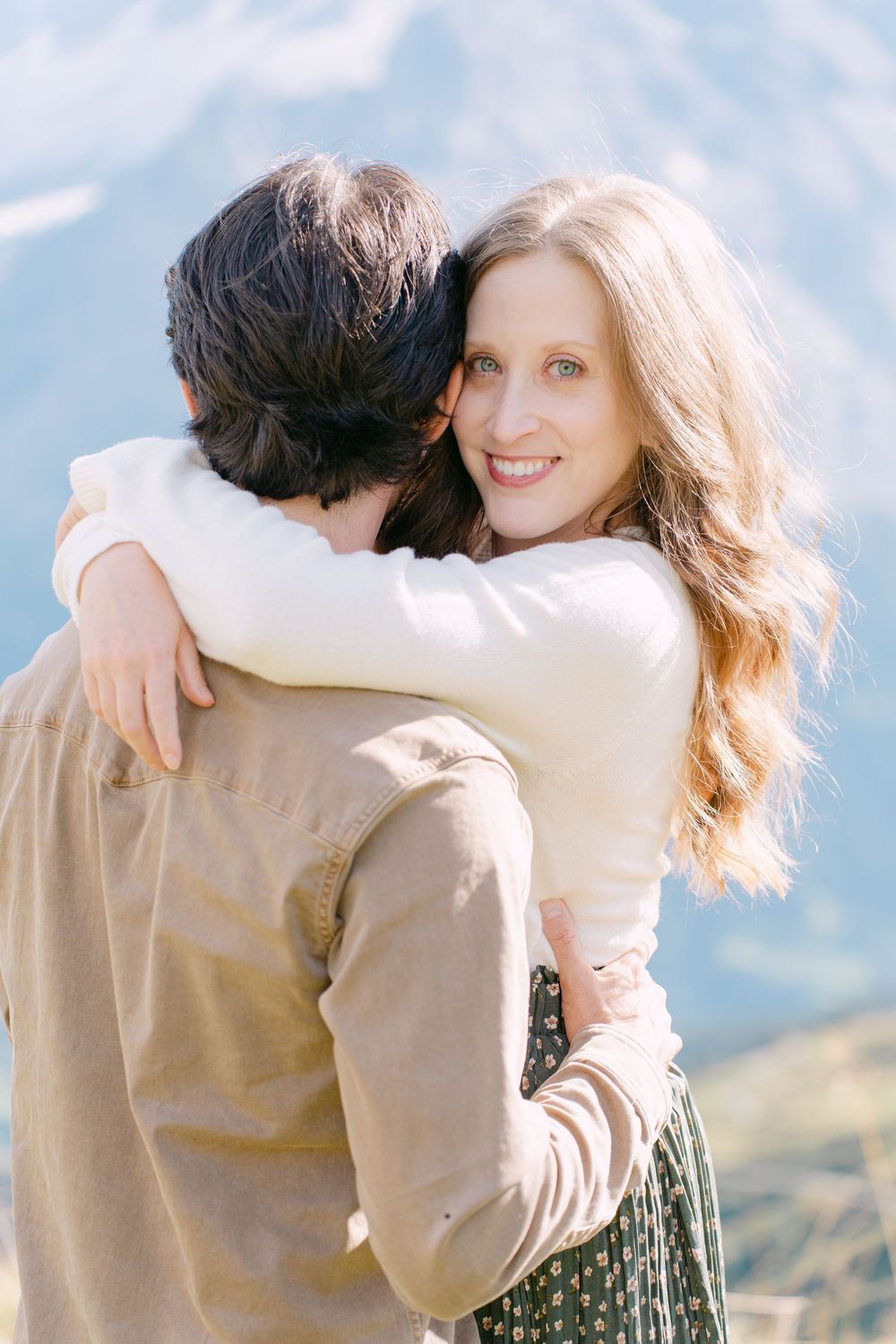 A romantic couple embraces outdoors against a scenic mountain backdrop on a sunny day.