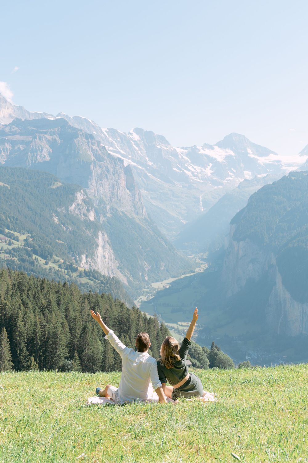 Two people sitting on grass with raised arms admiring stunning snow-capped Swiss Alps mountain vista in Lauterbrunnen Valley.