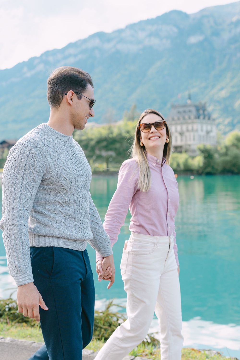 A couple in casual attire enjoys a romantic moment by a turquoise lake with mountains in the background.