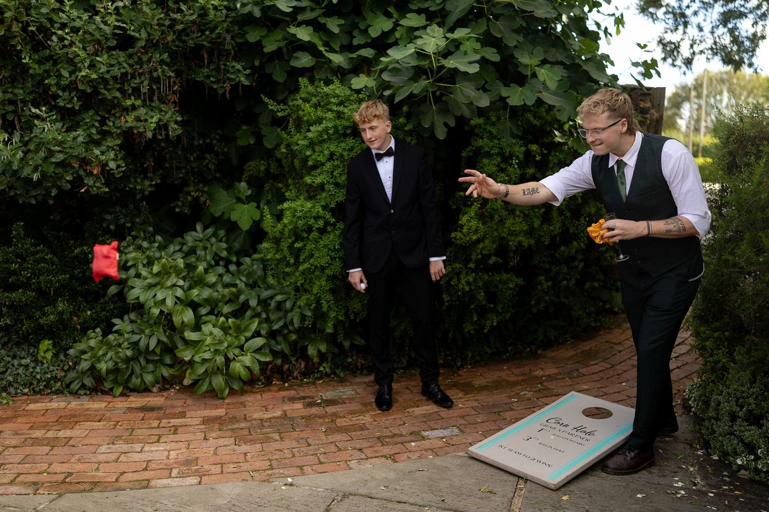 Two people in formal attire playing cornhole on a brick patio surrounded by green foliage.