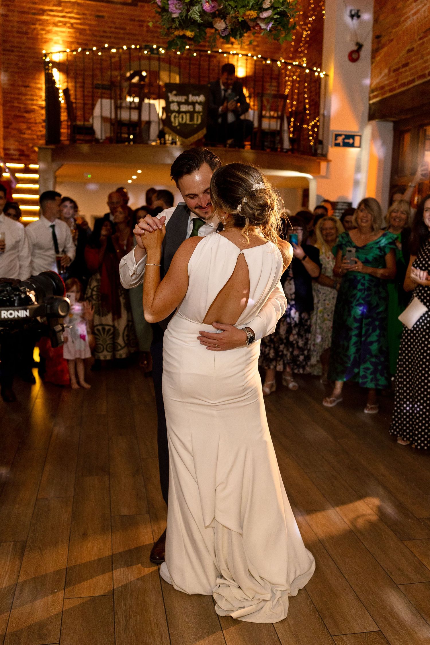 A couple shares their first dance at a wedding reception in a rustic venue with string lights and brick walls.