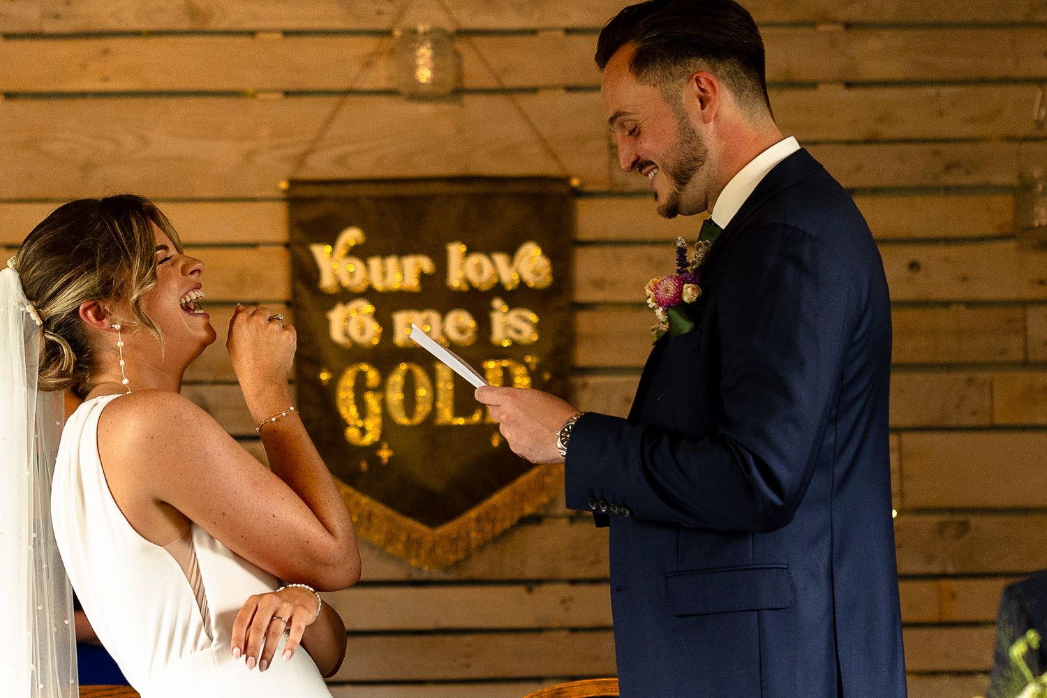 A couple shares a joyful moment at their wedding in front of a rustic wooden wall with gold lettering decor.