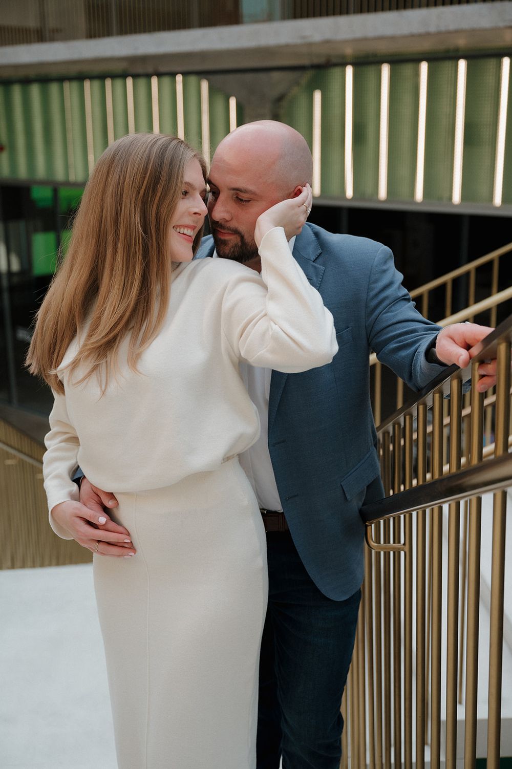 A romantic couple shares an intimate moment near golden railings in a modern architectural setting.
