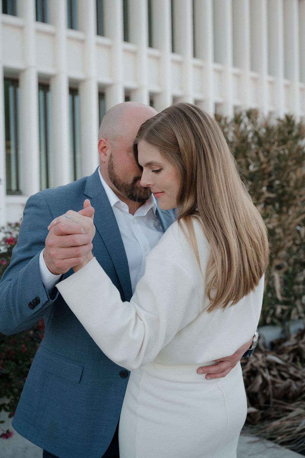 A couple in formal attire share an intimate dance moment outside a building with white columns.