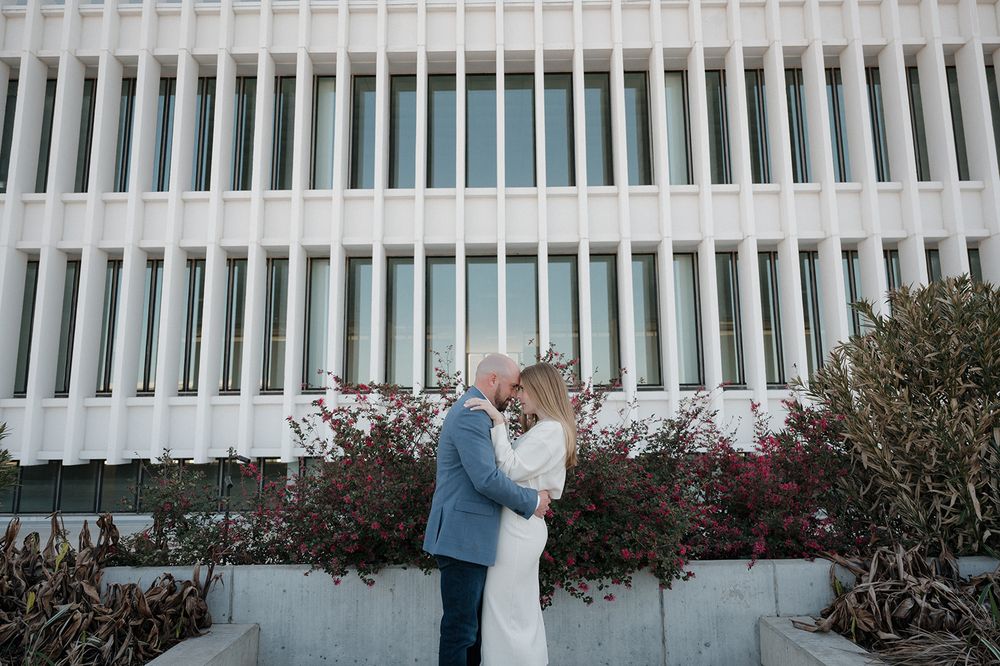 A couple embraces in front of a modern building with white vertical columns and flowering bushes.