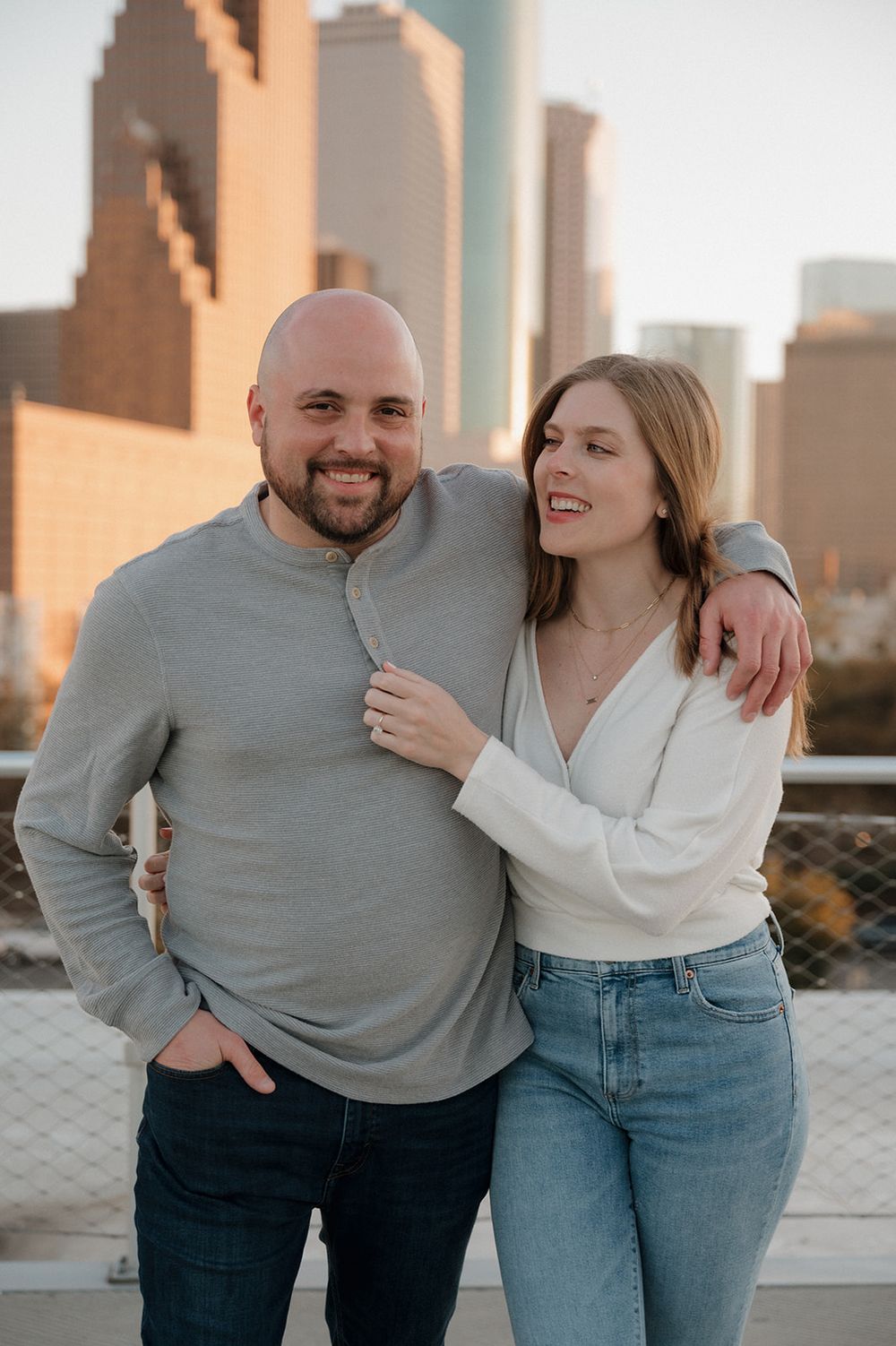 A couple embraces on an urban rooftop at sunset with city buildings in the background.