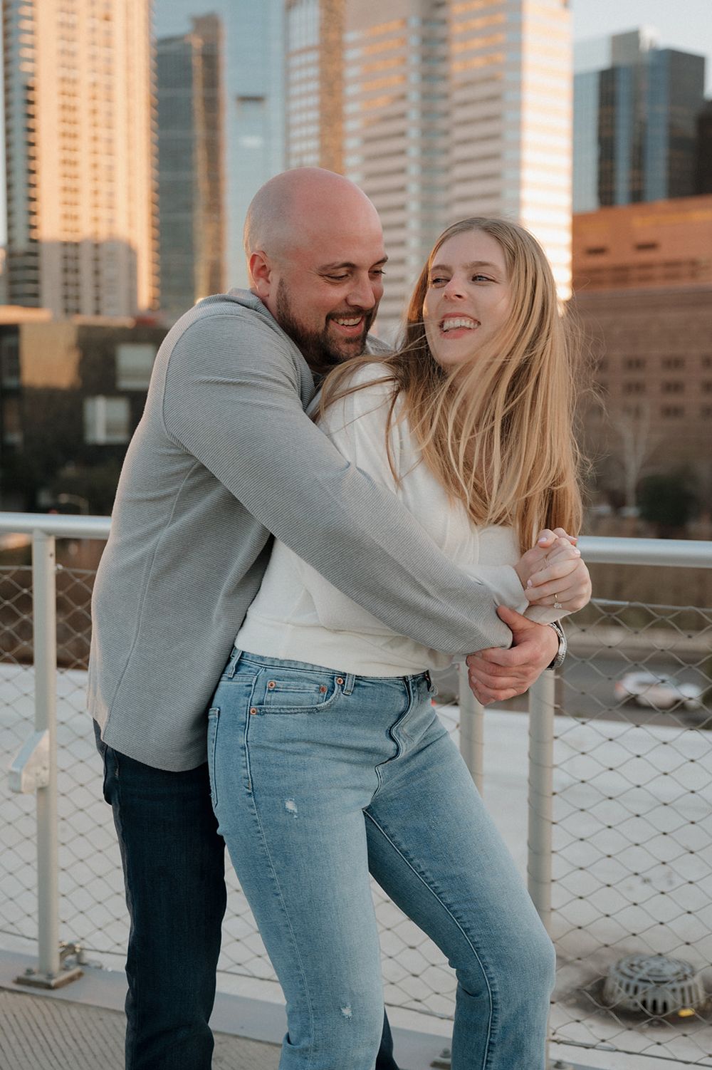 A loving couple shares a joyful embrace on a rooftop terrace with city buildings in the sunset background.