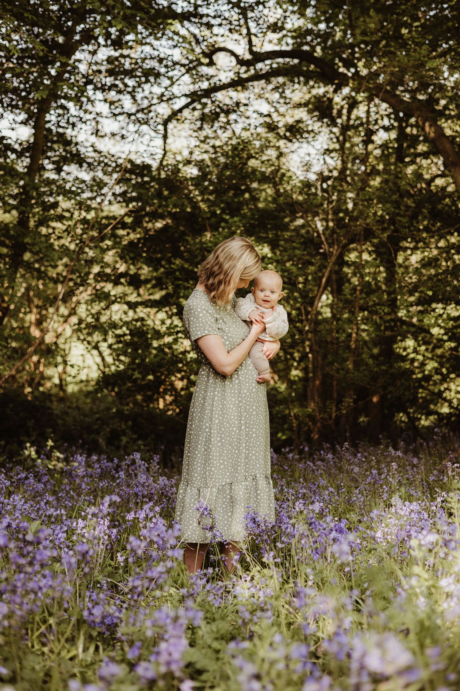 A figure in a polka dot dress stands amid a field of vibrant purple wildflowers at sunset in a wooded setting.