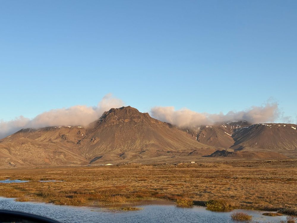A rugged mountain peak rises above brown marshland under a clear blue sky with wisps of clouds in Iceland.