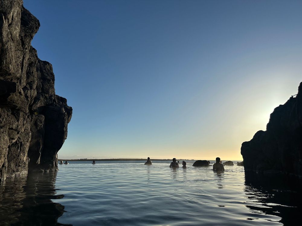 Silhouettes of kayakers paddle between rocky cliffs at sunset on calm waters.