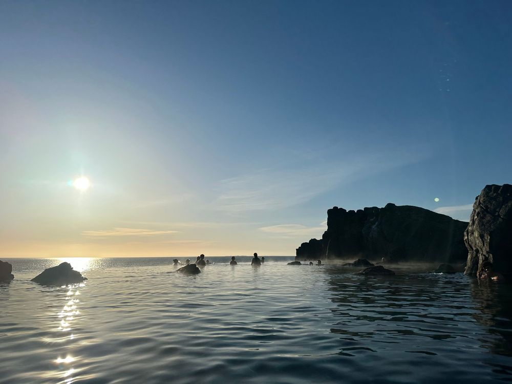 Silhouetted kayakers paddle through calm waters at sunset near dramatic rocky coastline formations.