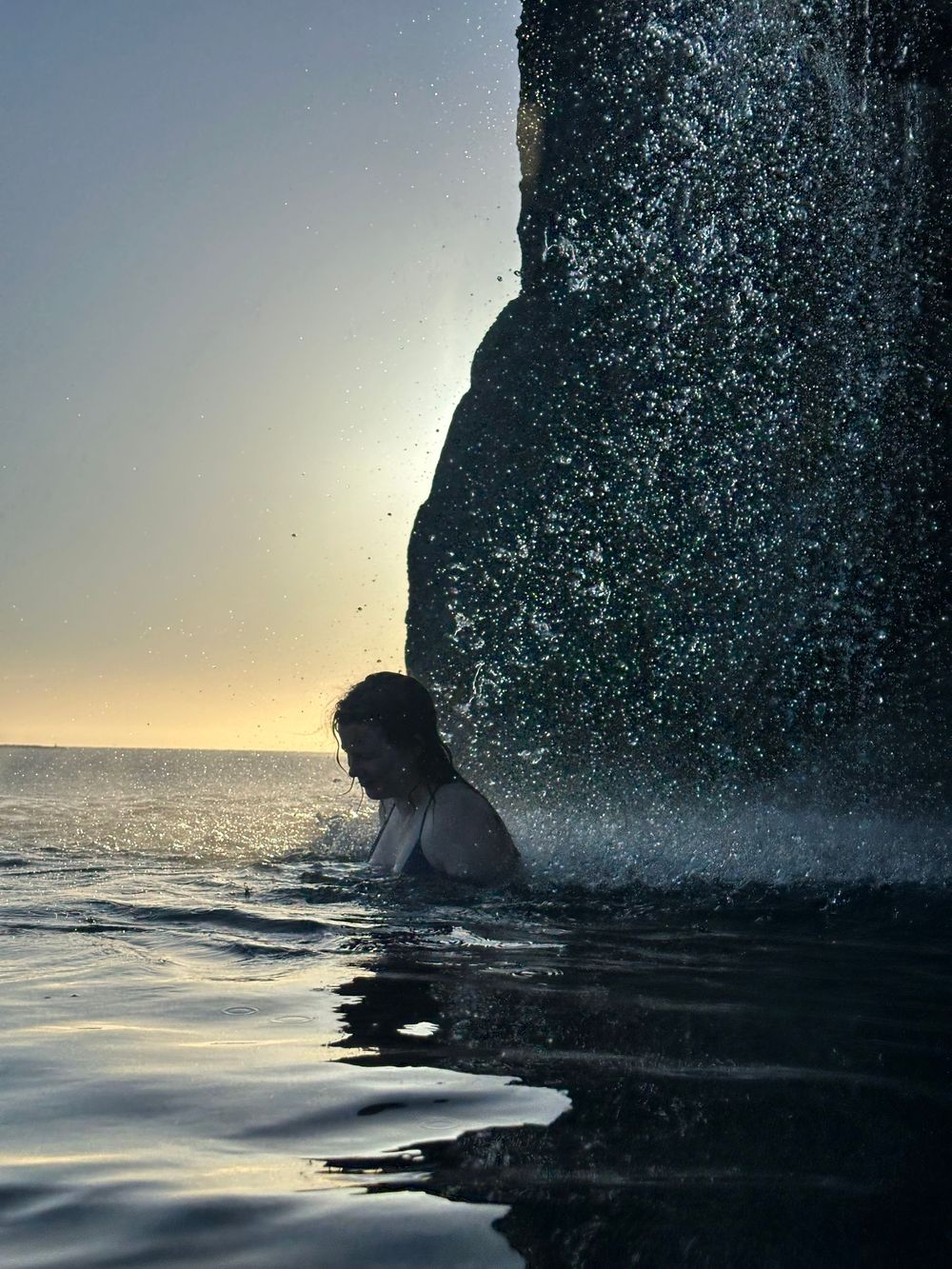 A silhouetted figure swims in the ocean at sunset near a towering cliff face with splashing water.