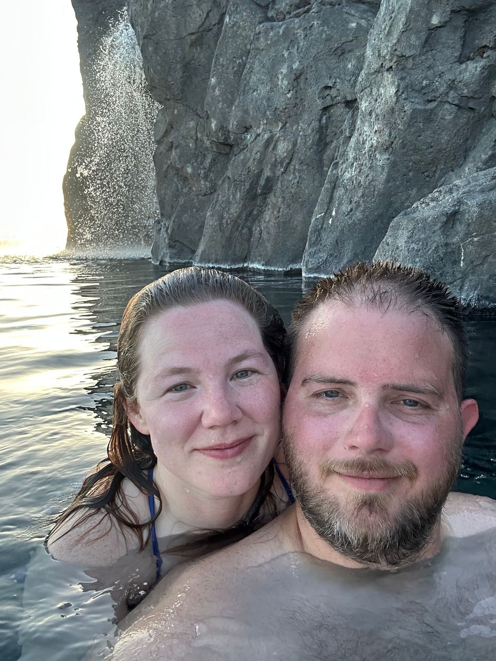 Two people taking a selfie while swimming near a tall cliff face with water and rocky formations at sunset.