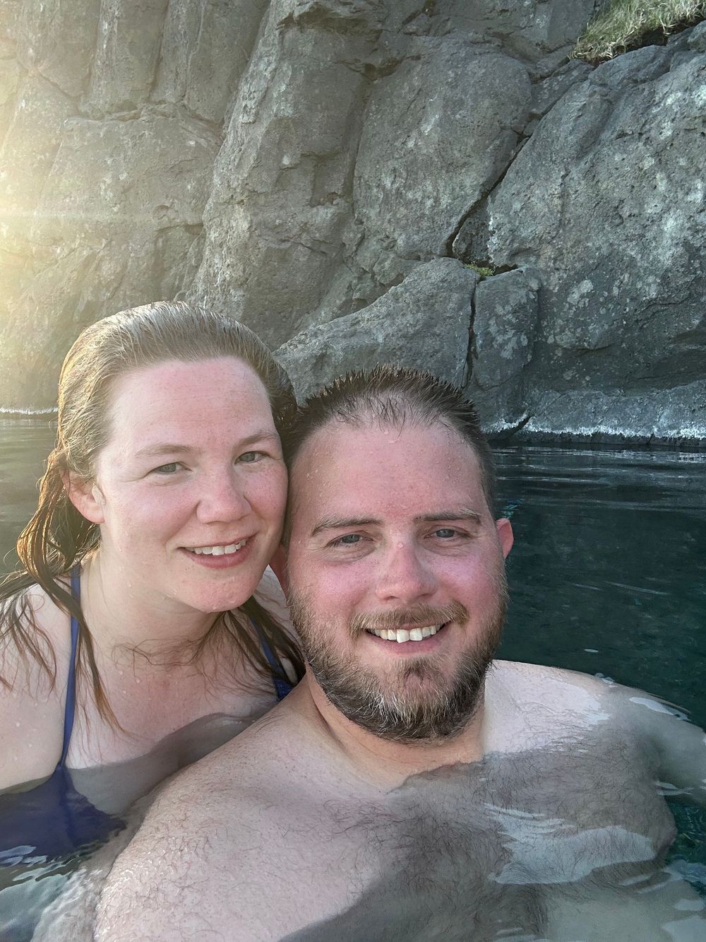 Two people enjoying a swim by rocky cliffs with sunlight streaming in the background.