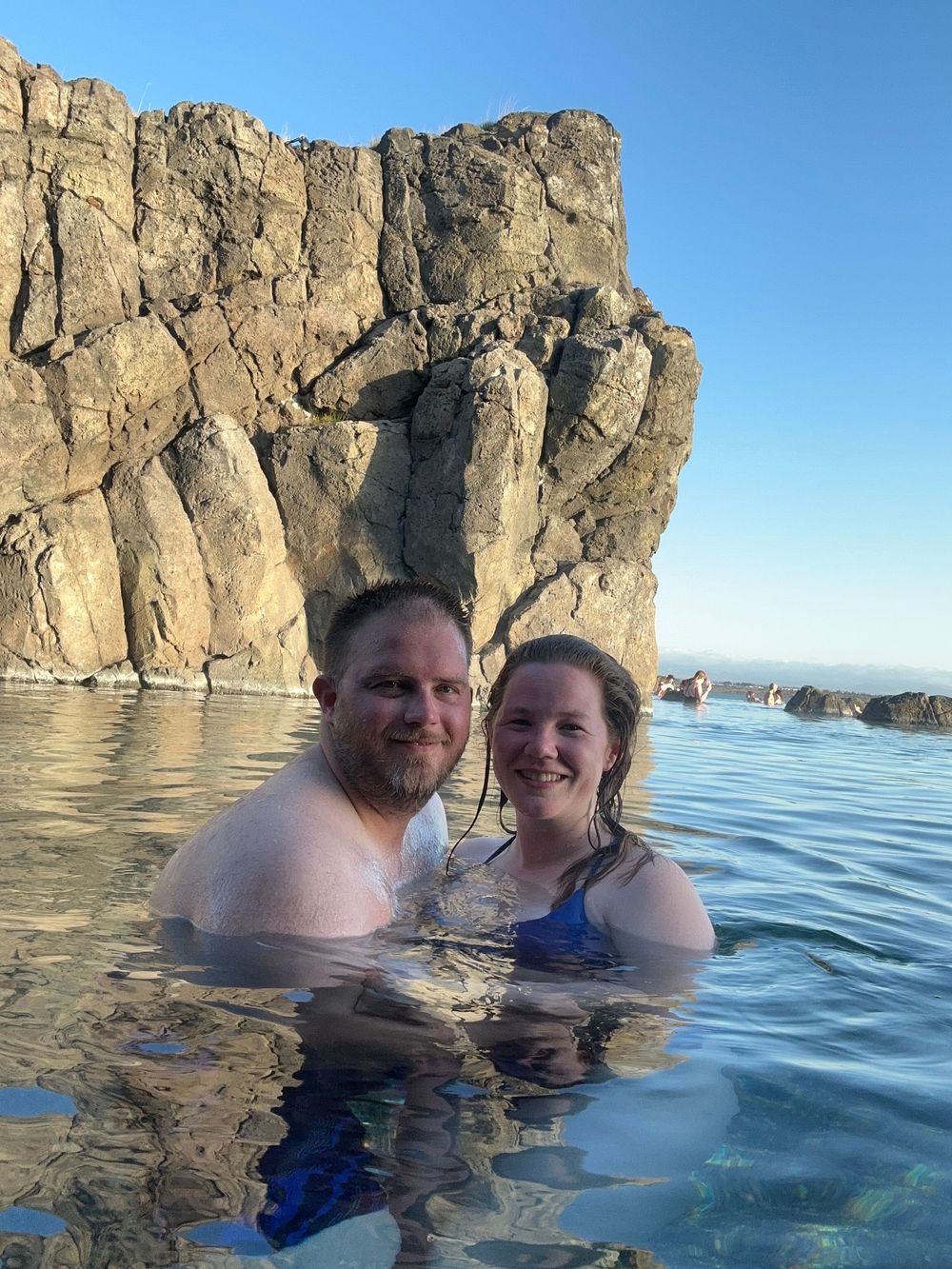 Two swimmers enjoying the crystal clear waters near a dramatic rocky cliff formation on a sunny day.