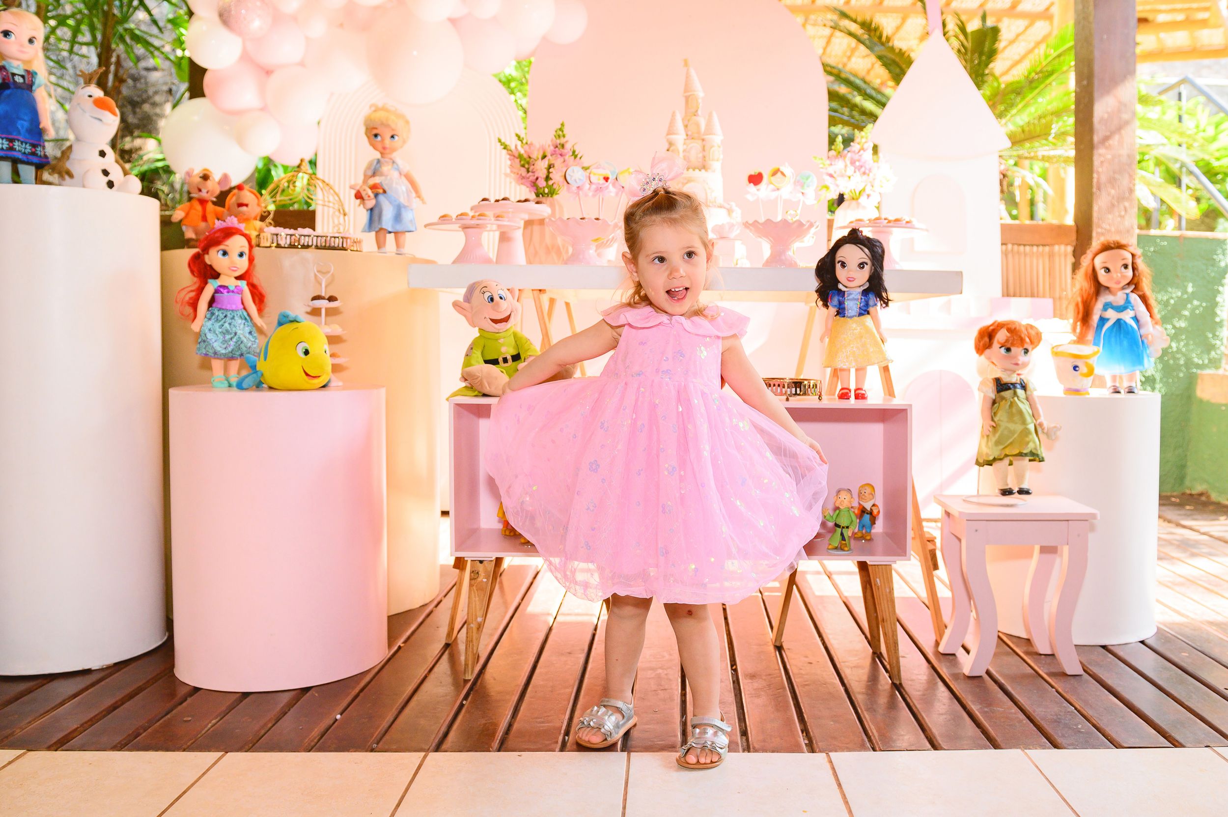 A playful pink dress twirls surrounded by Disney princess dolls displayed on pink pedestals on a wooden deck.