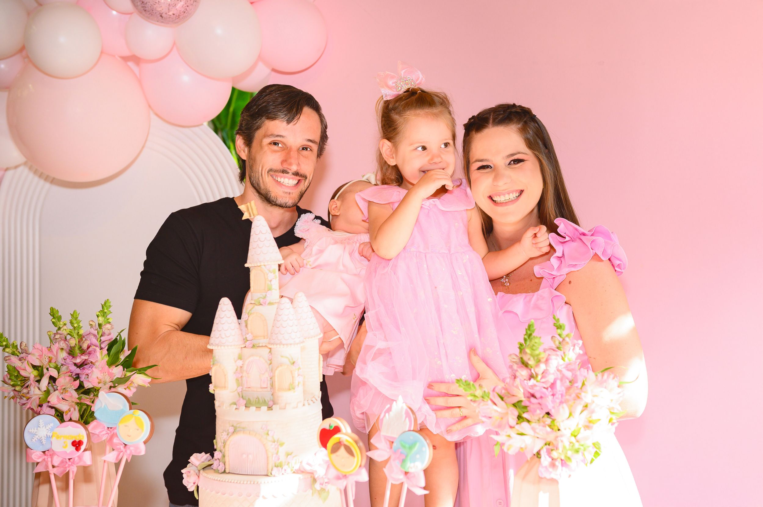 A family celebrates at a pink-themed party with decorative balloons, flowers, and a white cake.