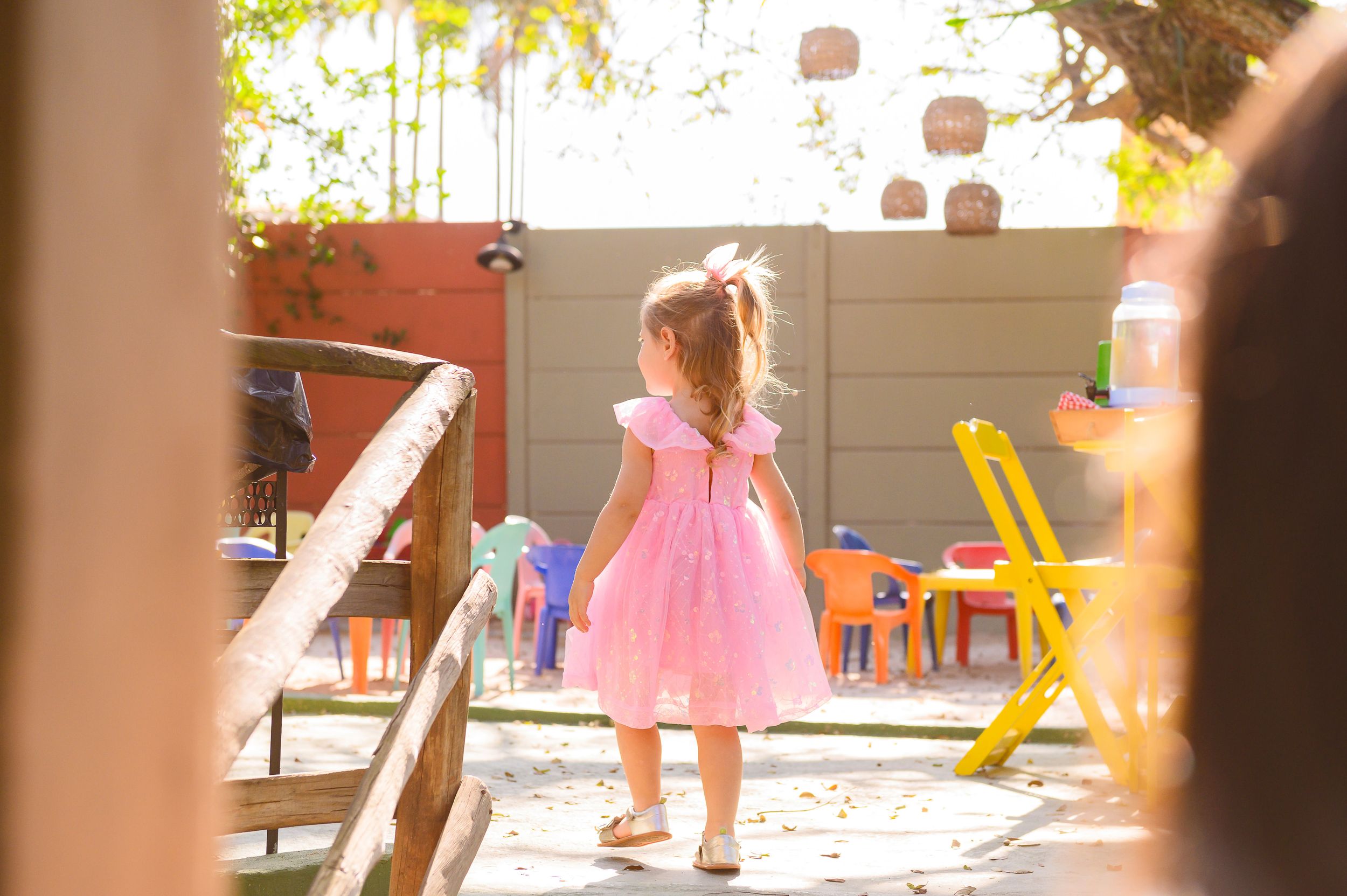 A small figure in a pink dress twirls and plays in a sunlit outdoor patio space with colorful furniture.