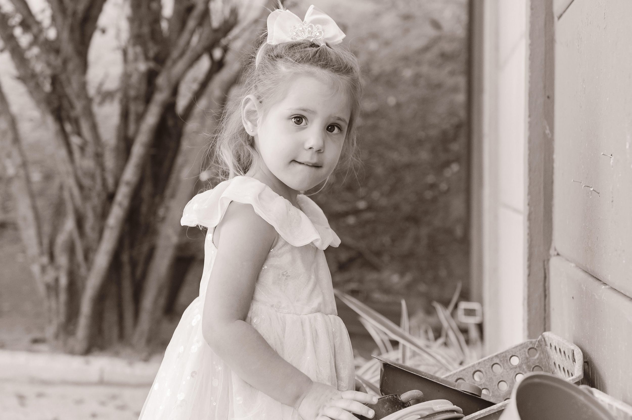 A vintage black and white photograph shows a young child in a white dress with hair bow standing near a column outdoors.