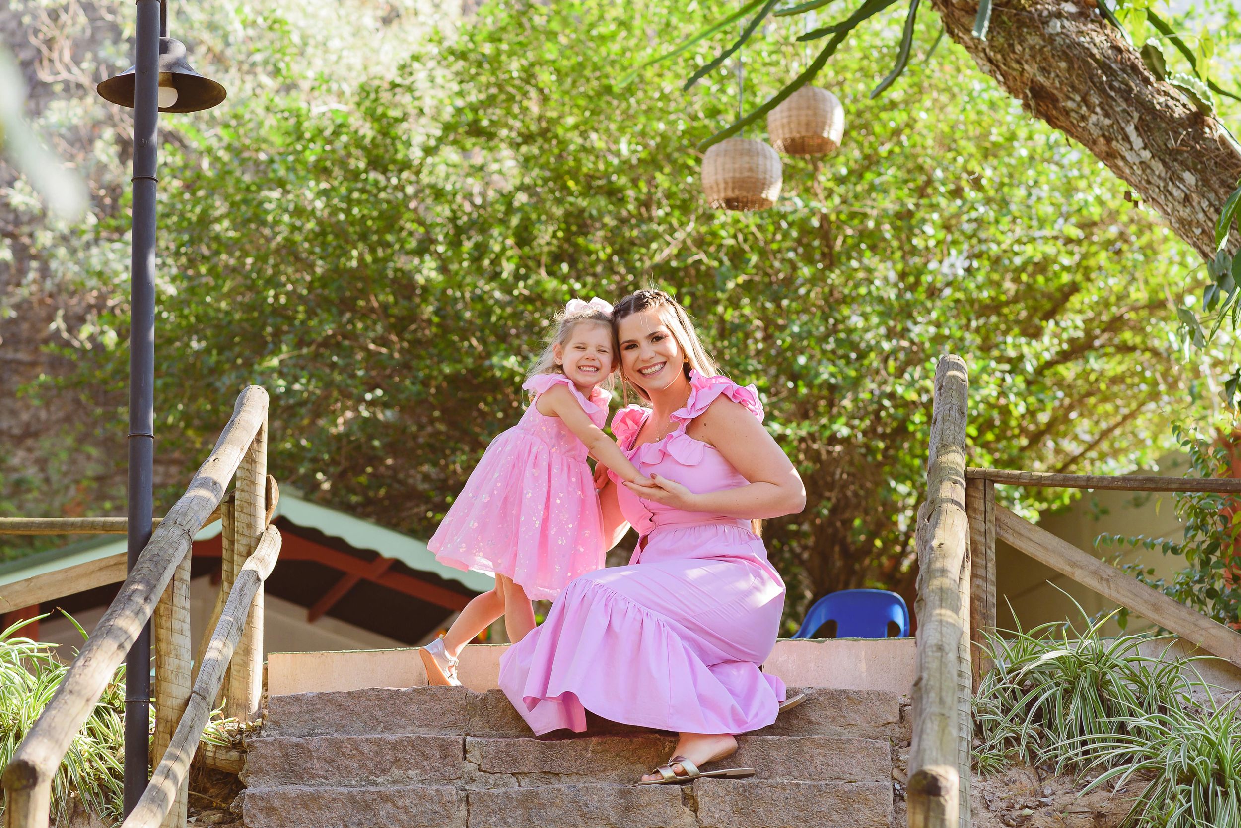 Two people in matching pink dresses sit together on a rustic wooden footbridge in a garden setting.