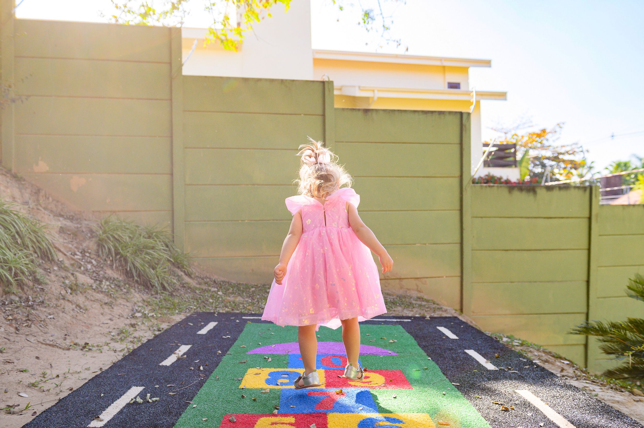 A child plays on a colorful hopscotch path wearing a pink dress in bright sunlight against a green wall.