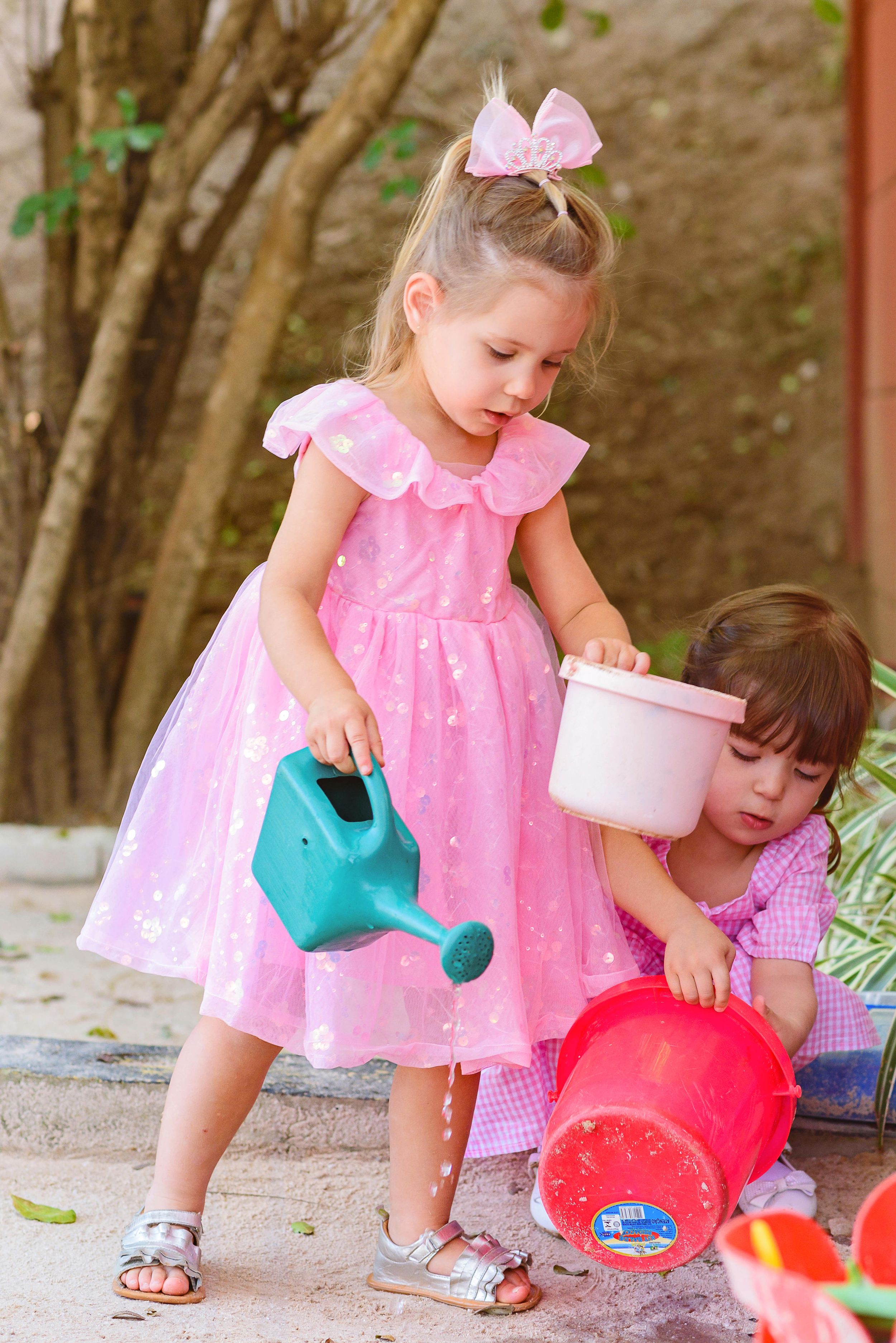 Two young children play together outdoors with colorful watering cans and buckets on a sunny day.
