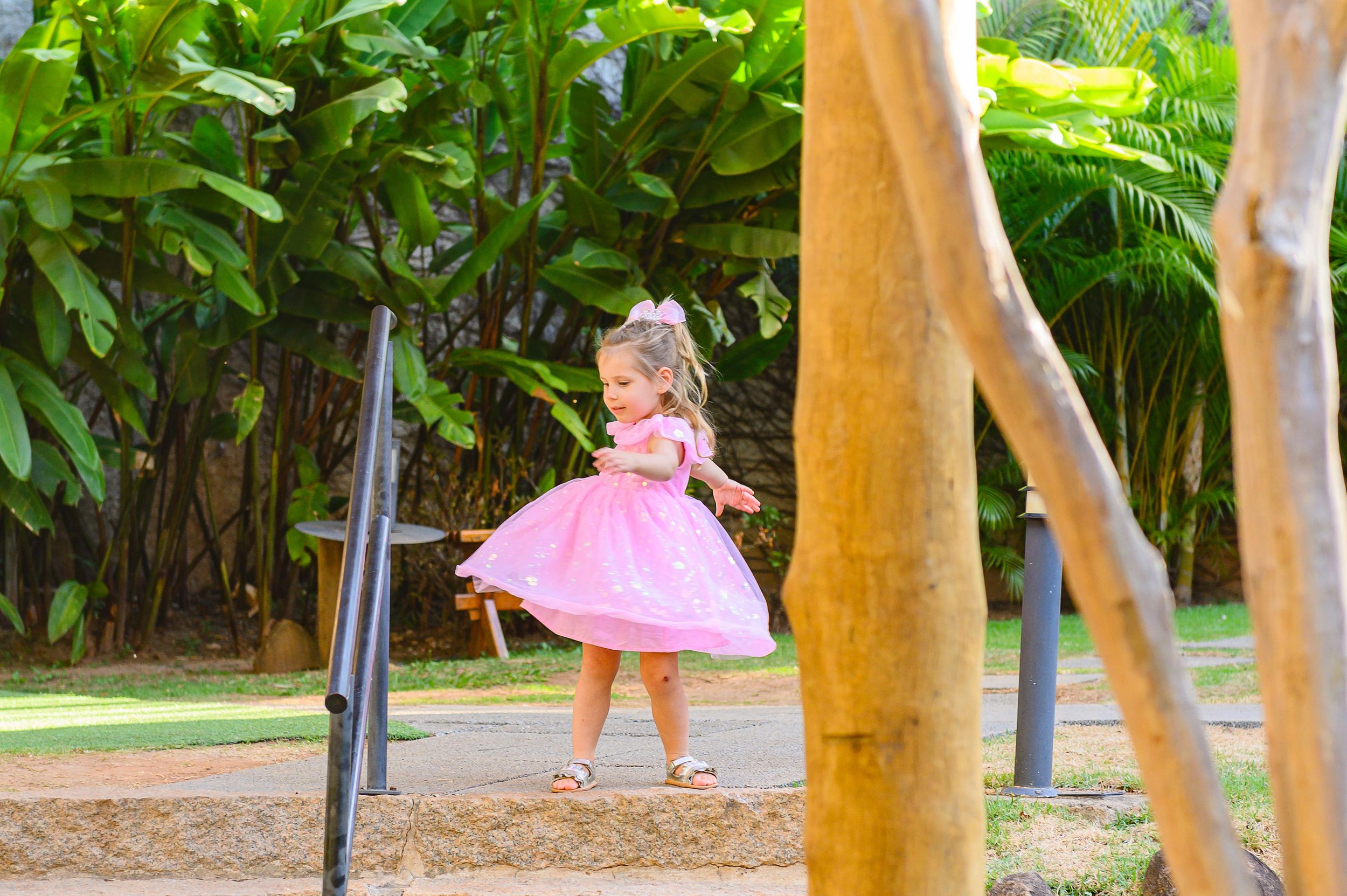 Small figure in a pink dress spins gracefully in a tropical garden setting with palm trees and lush greenery.