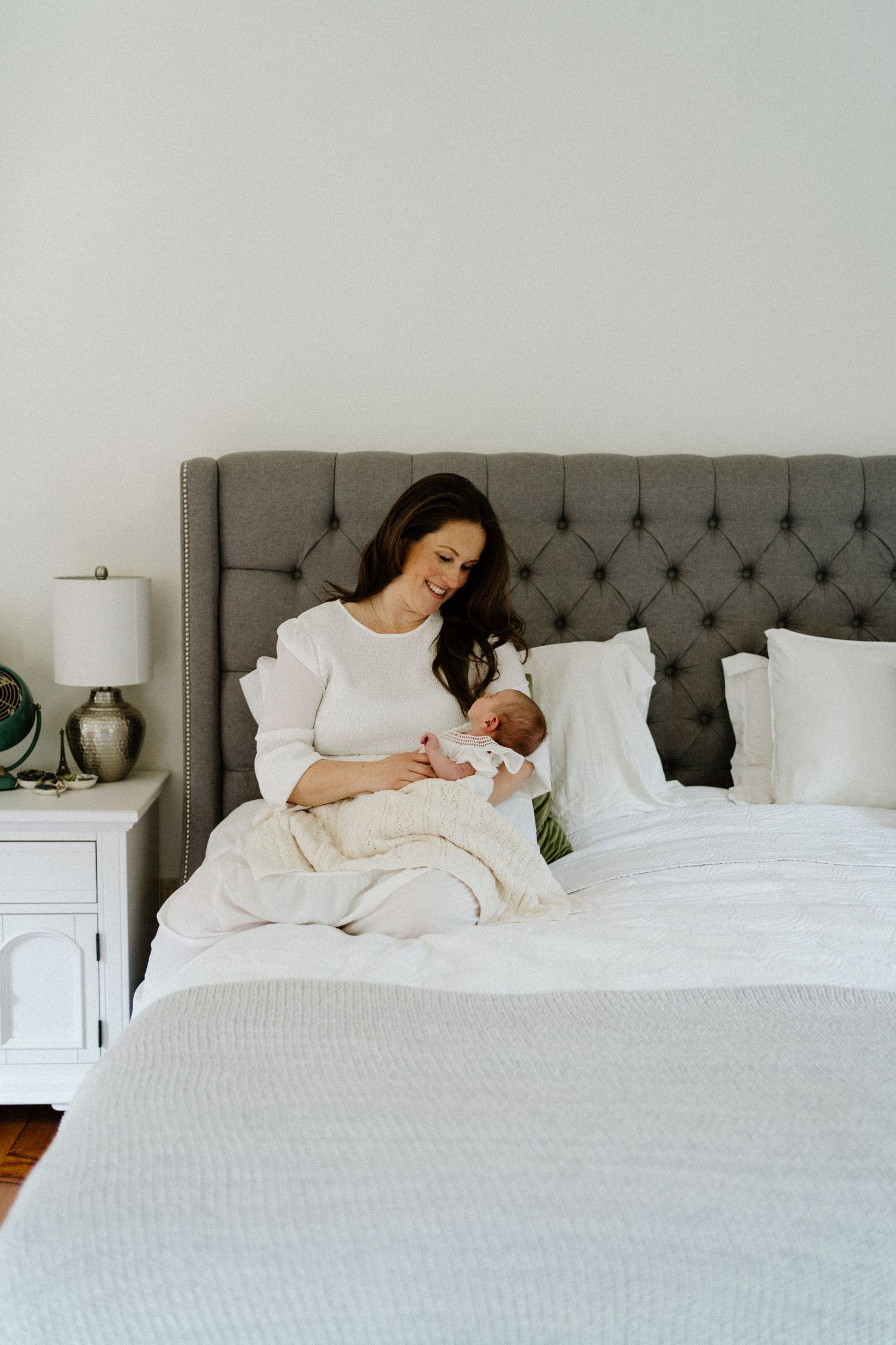 A person in white sits on a grey tufted headboard bed while cradling a baby in a bright, minimal bedroom setting- Muskoka newborn session