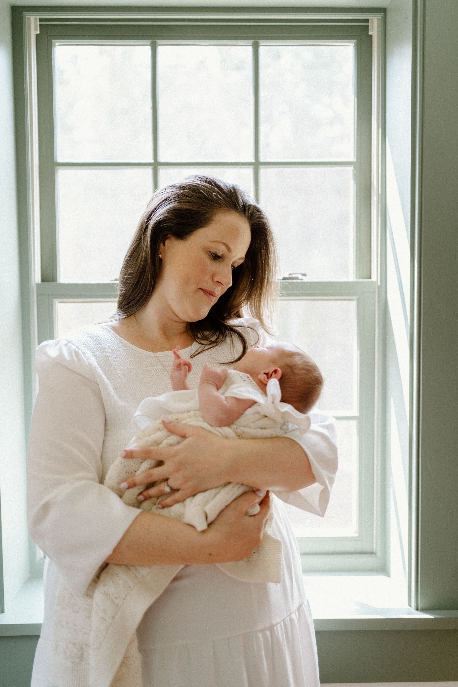 A tender moment by a bright window as a parent cradles a newborn baby wrapped in white.
