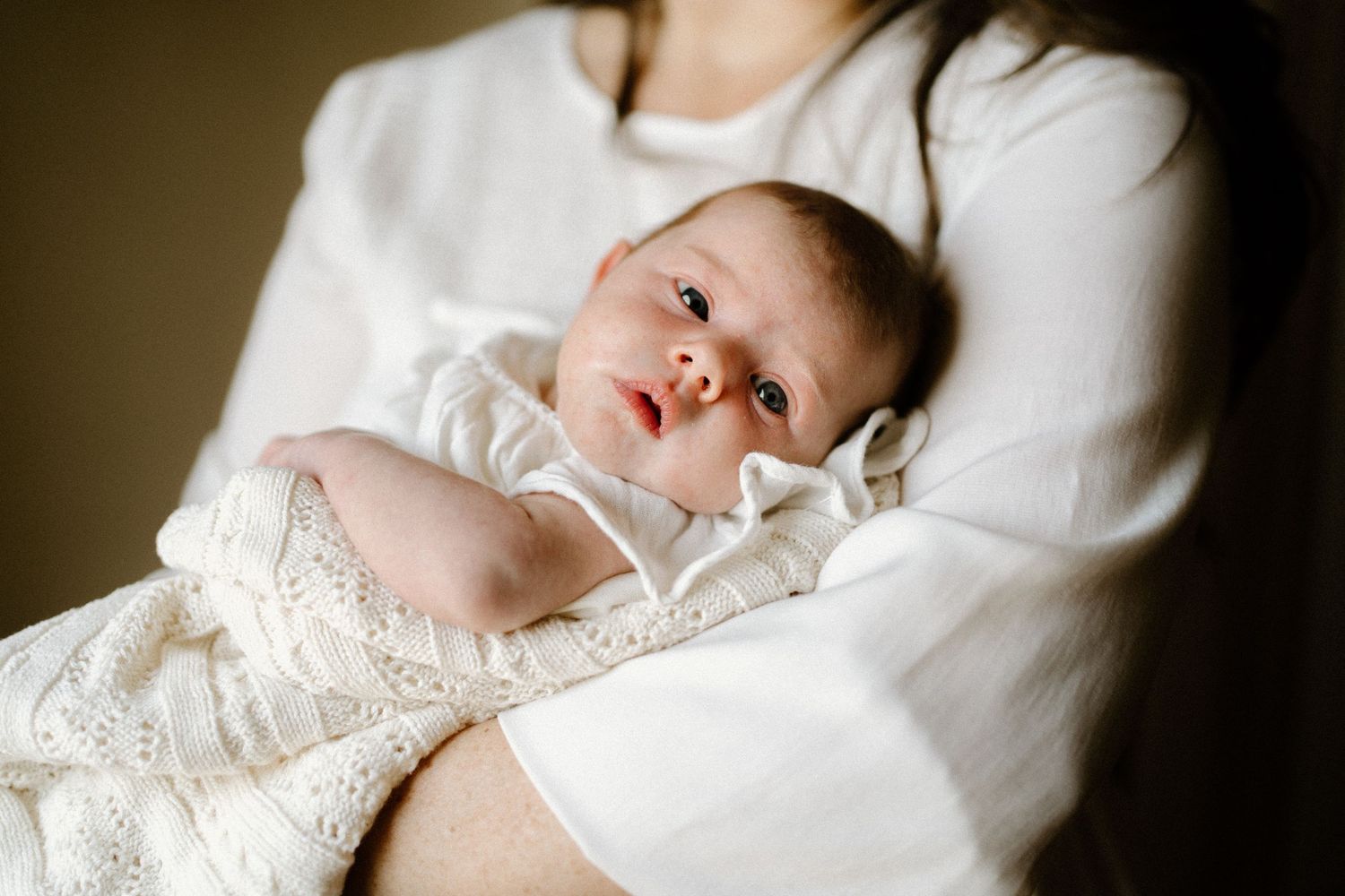 A small infant wearing a delicate white lace christening gown rests in a caregiver's lap.