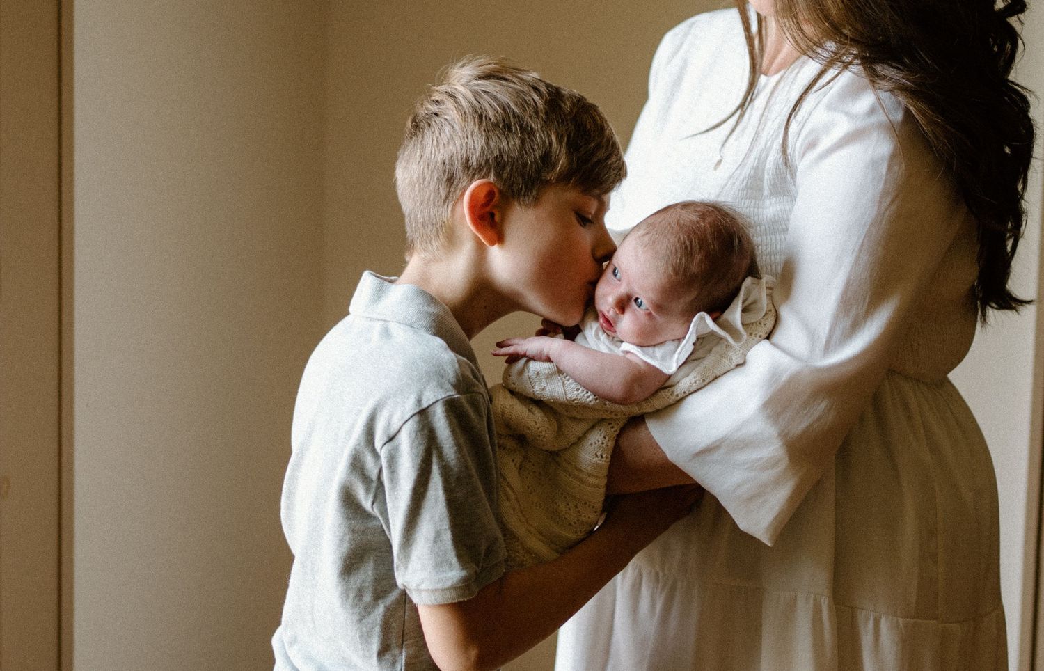 Sweet bonding moment between young boy and newborn baby as mother stands nearby in white dress by window.
