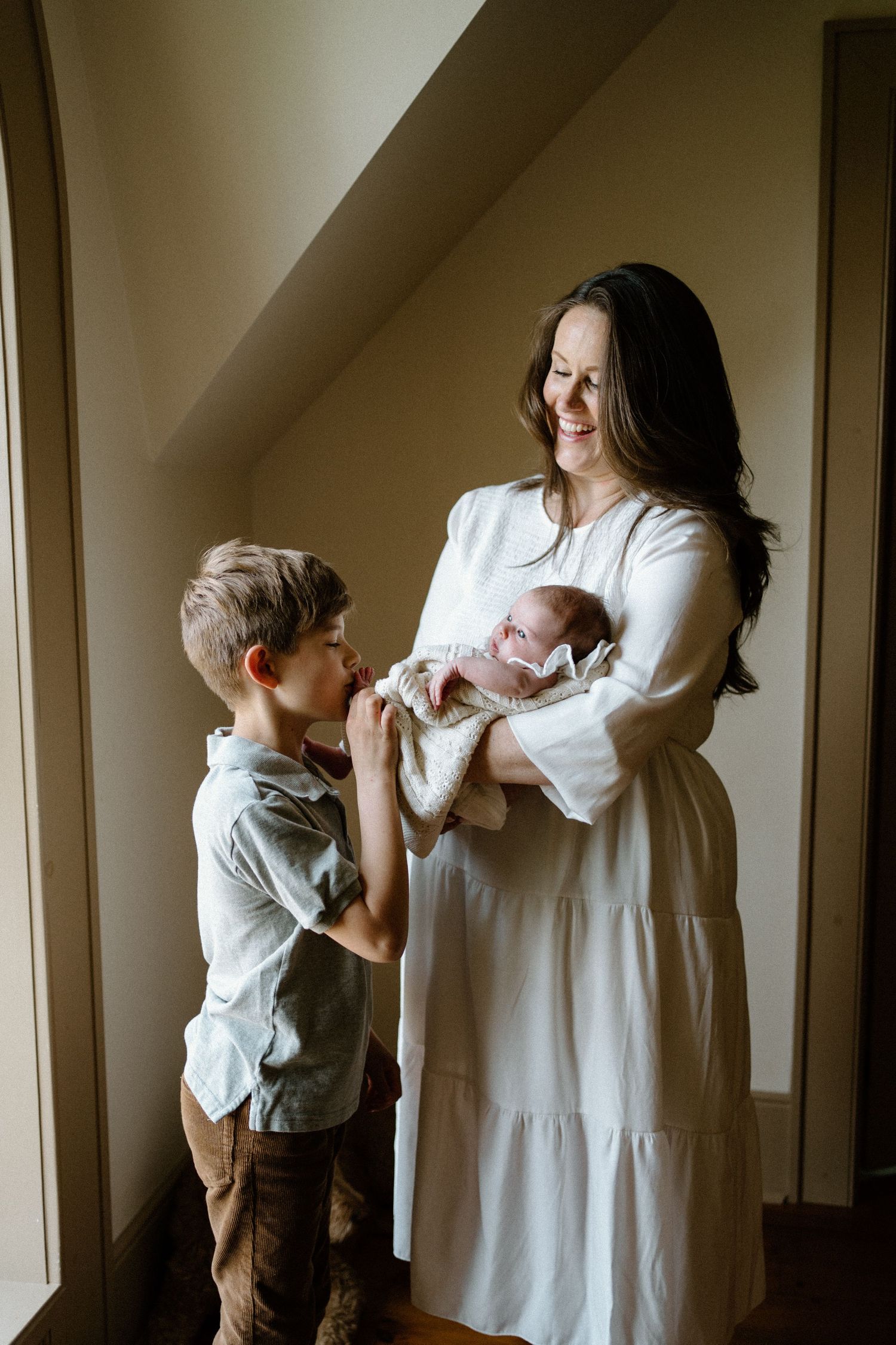 A touching family moment as a sibling in brown pants meets their new baby by a window.