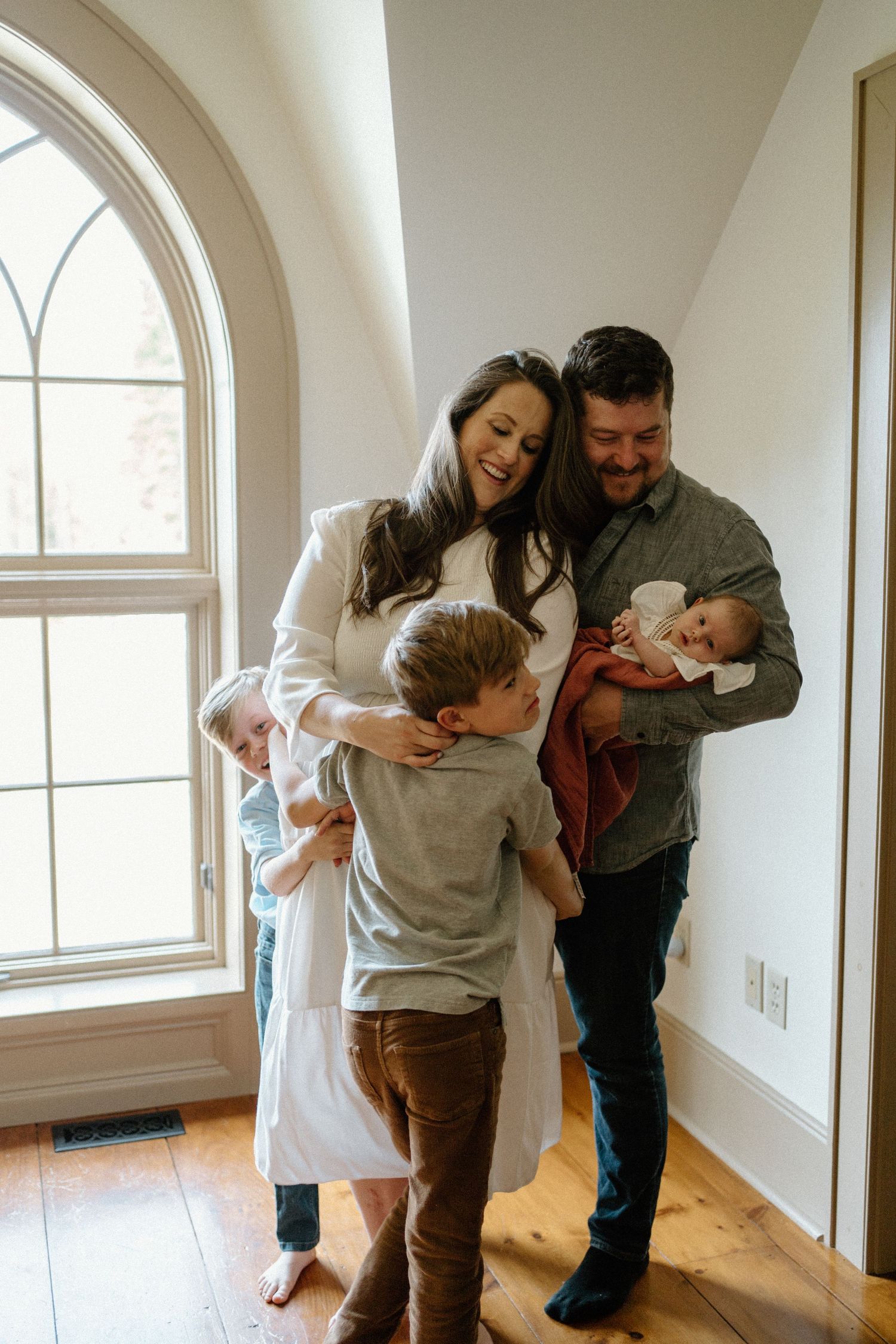 Family shares tender moment together by arched window in bright sunlit room with hardwood floors.