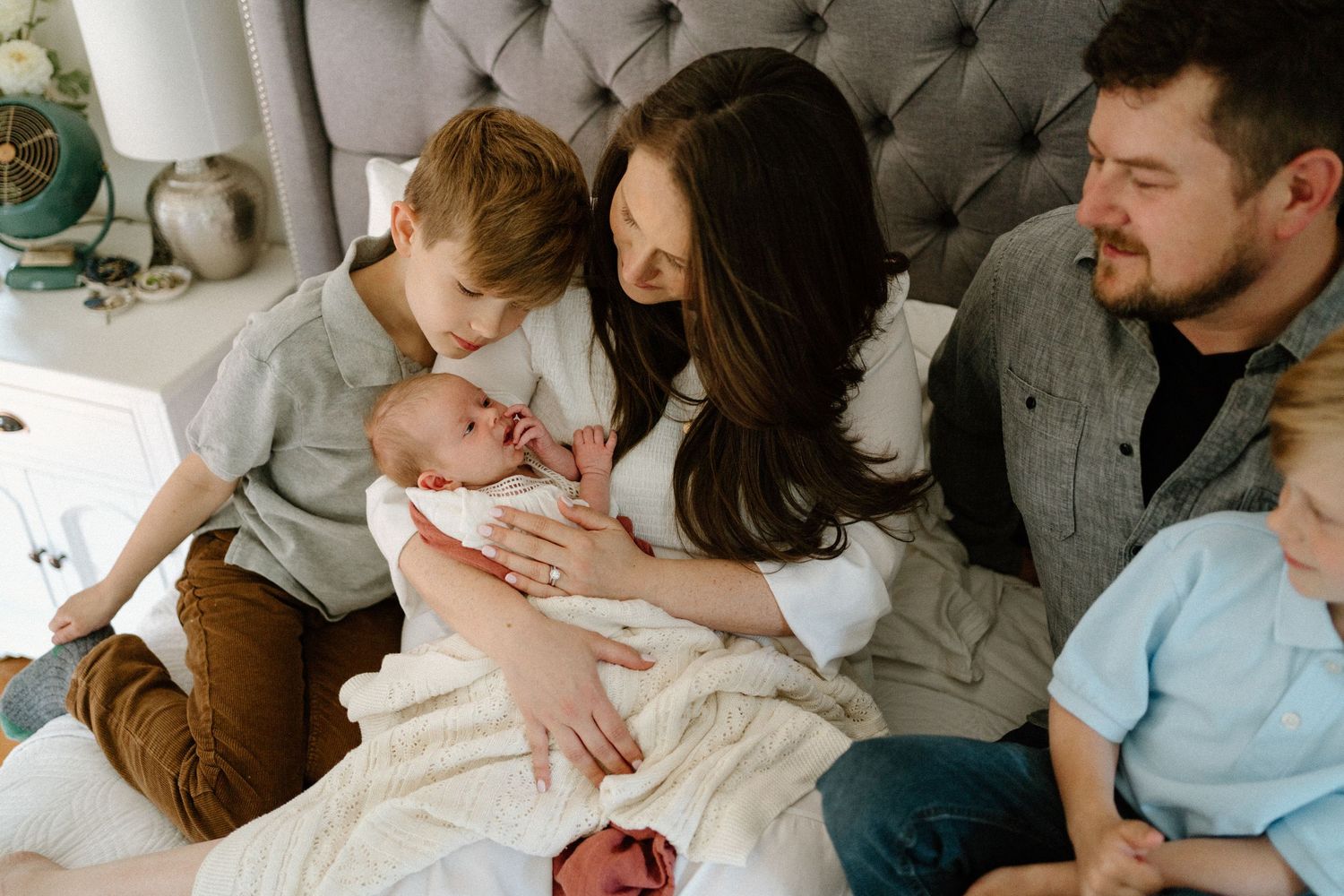 A family relaxes together on a gray tufted headboard bed while tenderly embracing their newborn baby.